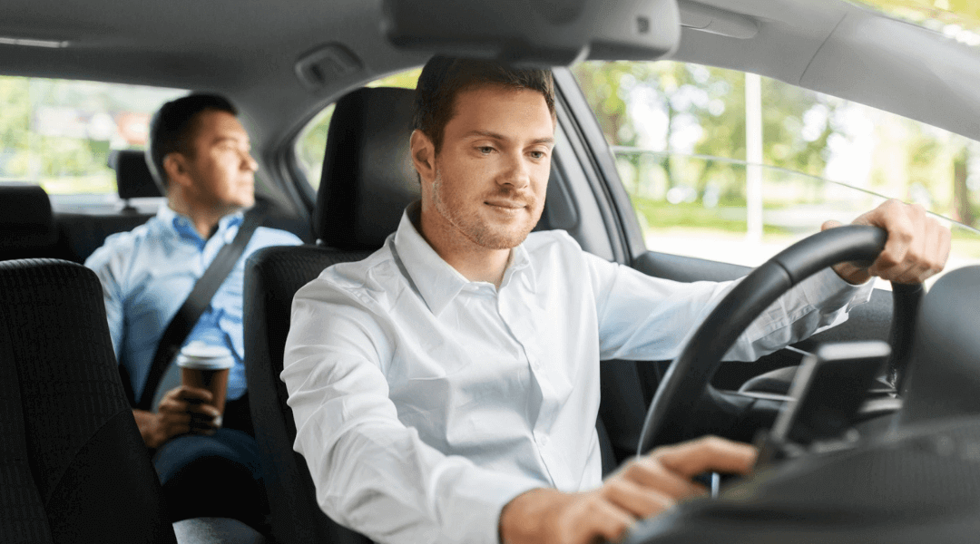 Inside a car, one man is driving while the other is seated beside him, both focused on the road ahead.