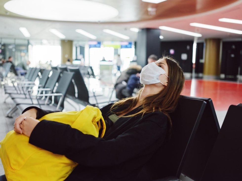 A woman in a face mask sits on a chair at an airport, waiting for her flight amidst a busy terminal environment.