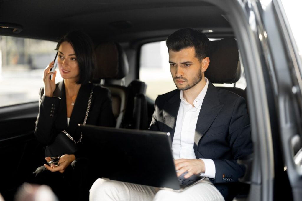 Inside a car, a man and woman collaborate on a laptop, engaged in their work.