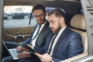 Two businessmen in suits seated in the back of a car, engaged in conversation.