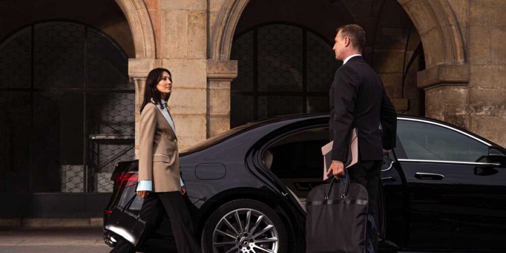 A chauffeur holding a bag opens the door of a black luxury car for a woman outside a stone building