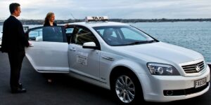 A man in a suit opens the door of a white corporate taxi for a woman by the water