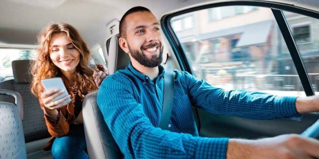 A smiling bearded man is driving a car while a woman in the back seat looks at her smartphone