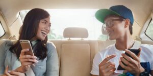 A man and woman sit in a car's back seat, both focused on their cell phones.