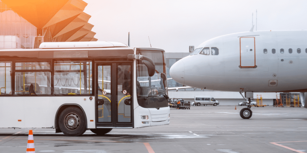 A white airport bus is parked on the tarmac near a large passenger airplane. The scene is bright with an orange hue above, indicating a sunset.