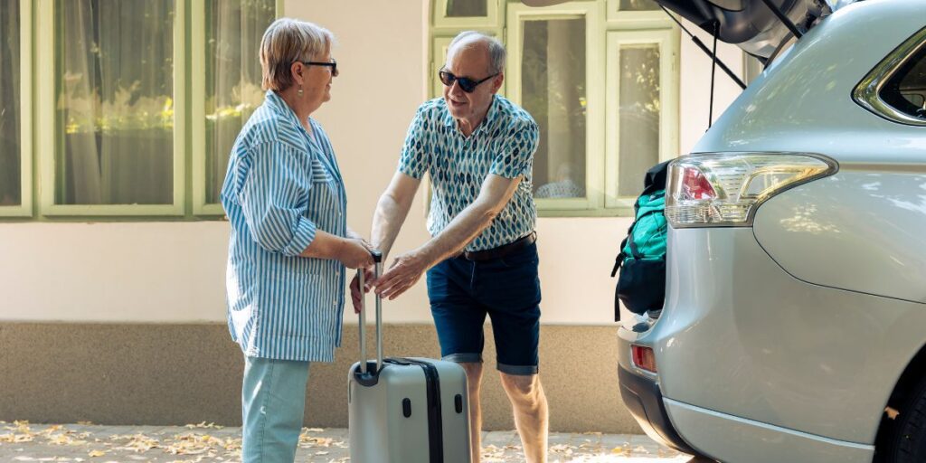 A senior couple standing next to an open car trunk while handling a silver suitcase on a sunny day outdoors.