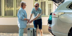 A senior couple standing next to an open car trunk while handling a silver suitcase on a sunny day outdoors.