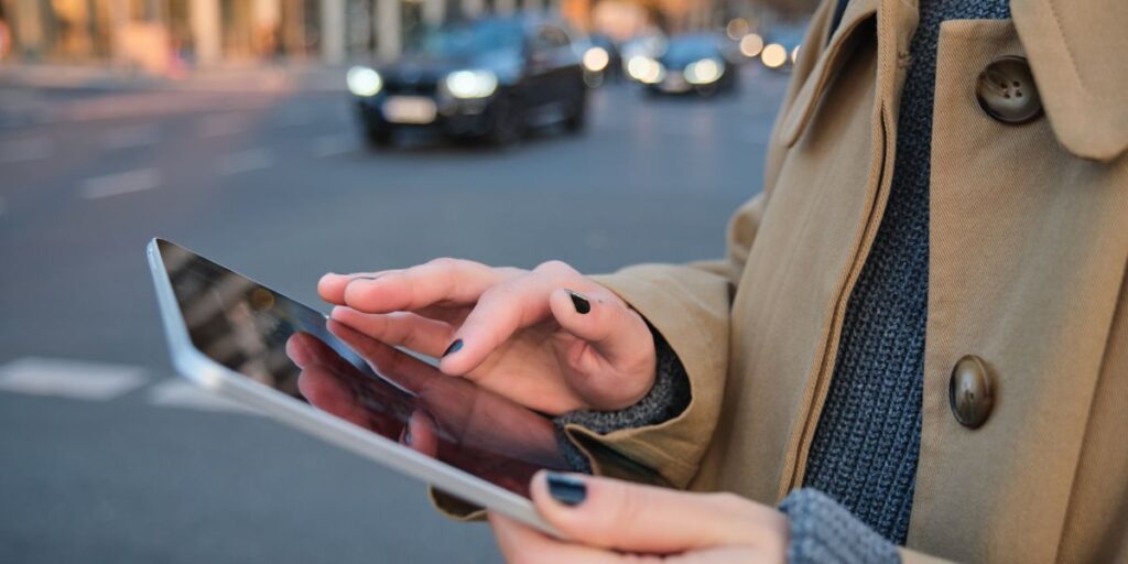 Close-up of a person's hands with dark nail polish using a digital tablet outdoors near a road with traffic
