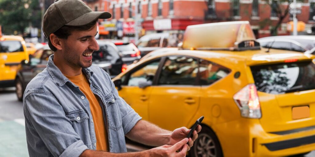 A smiling man in a cap uses his smartphone on a busy city street with yellow taxis blurred in the background.