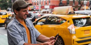A smiling man in a cap uses his smartphone on a busy city street with yellow taxis blurred in the background.