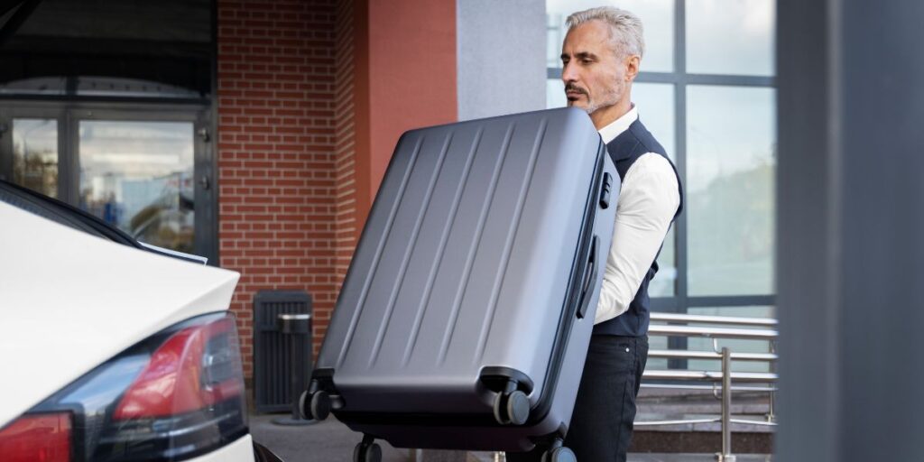 A professional driver in a vest lifts a large silver suitcase to load it into the trunk of a white car
