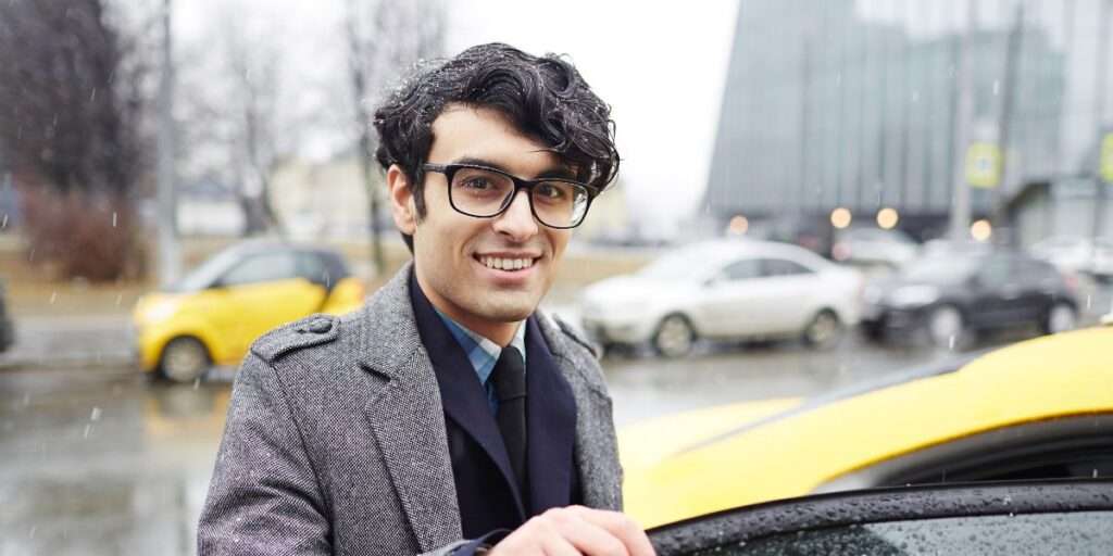 A smiling man in a grey coat and glasses standing outdoors on a rainy day next to a bright yellow taxi cab