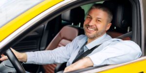 A friendly taxi driver smiling while sitting behind the wheel of a yellow car with his seatbelt securely fastened