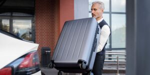 A man in a vest carefully lifting a large grey hard-shell suitcase to load it into the trunk of a white vehicle.