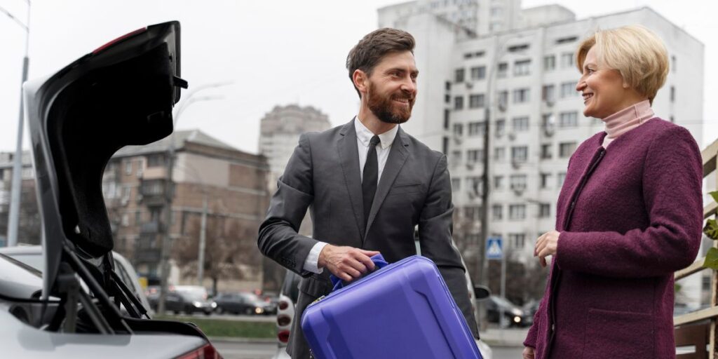 A smiling driver in a suit holding a purple suitcase while talking to a female passenger by an open car trunk.