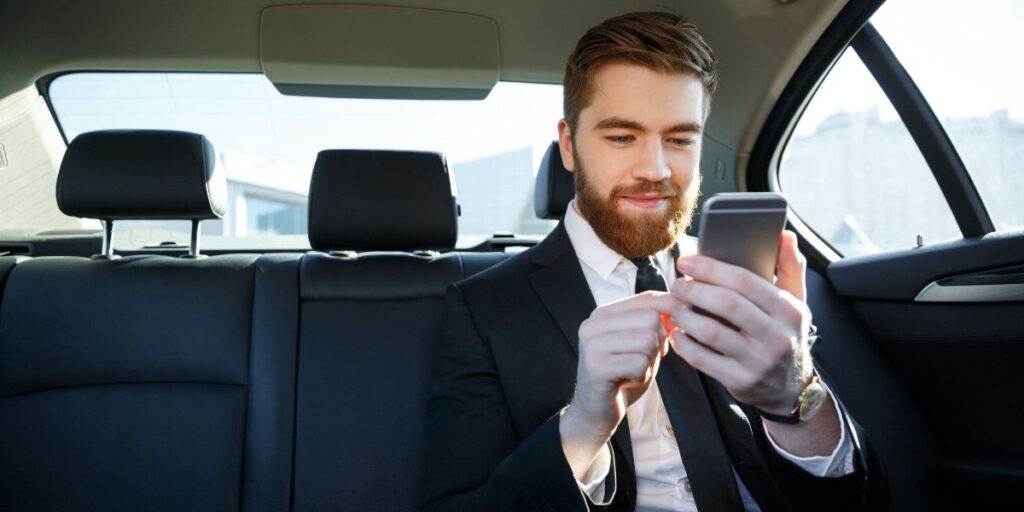A smiling businessman with a beard sitting in the backseat of a car and using his smartphone.