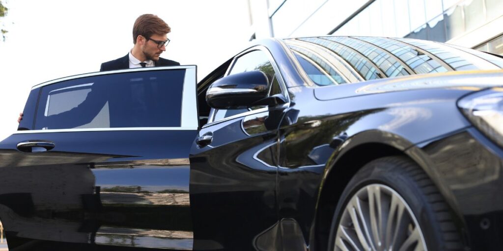 A professional chauffeur in a suit and glasses holding open the door of a luxury black sedan.