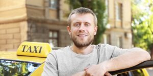 A smiling male taxi driver with a beard leaning against his yellow cab on a sunny street
